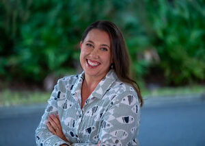 Smiling woman with crossed arms outdoors in front of green foliage.