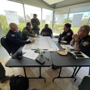 Individuals in different agency uniforms sitting at a table talking with each other over papers on the table working to develop the Binational Debris-Related Flooding Response Guide