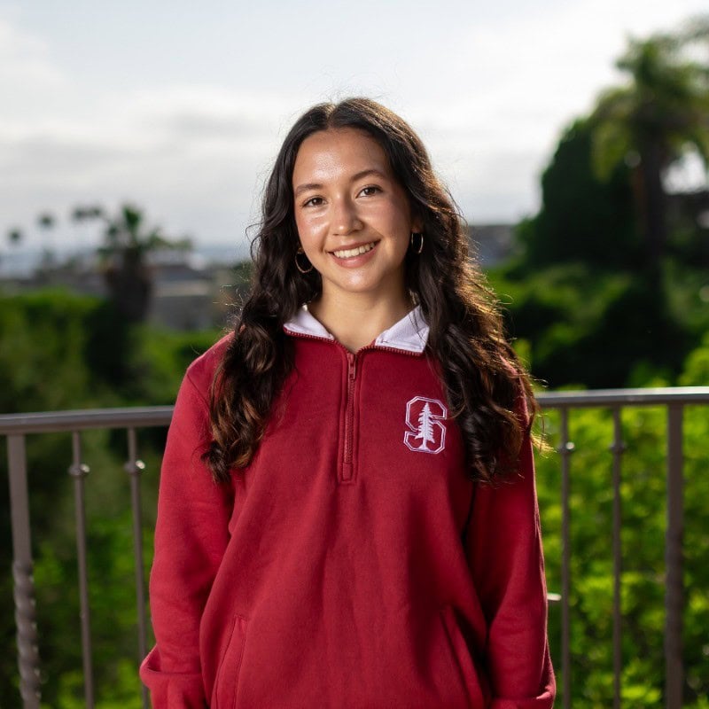 Young woman smiling outdoors with greenery and cloudy sky in background.